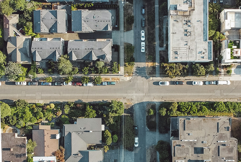 Aerial view of a residential neighborhood with houses, trees, and parked cars along a road.